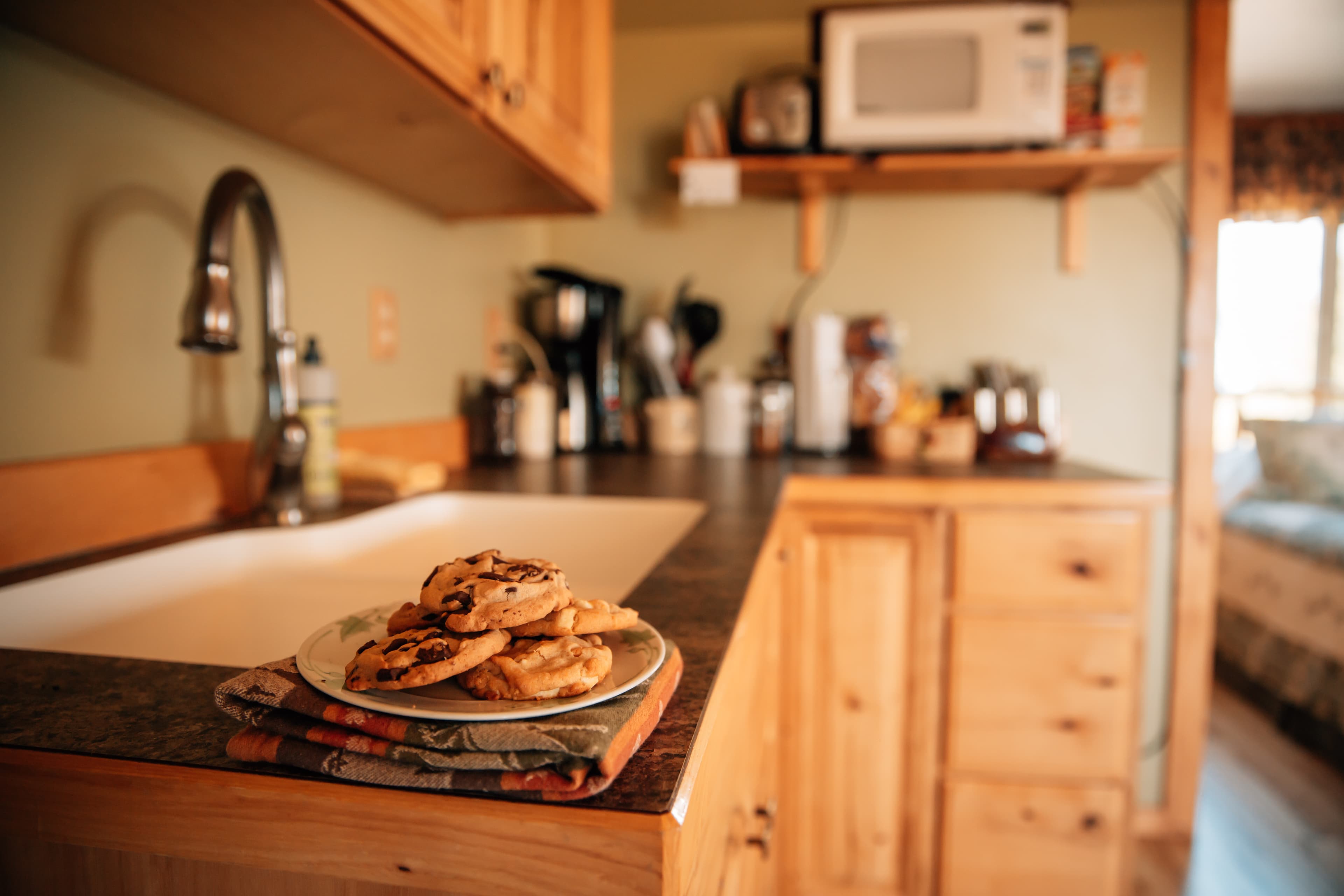 Beautifully finished wood kitchen counter and cabinets with sink, coffee maker, and plate of cookies, separate shelf with microwave